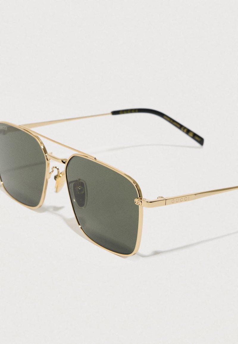 Gouden metalen zonnebril met vierkante lenzen, donker groene tint en slanke monturen. Bevat logo-accenten op de armen en heeft een minimalistisch ontwerp.