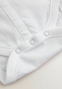 Beli bombažni bodi z teksturiranim, mrežastim vzorcem. Ima okrogel ovratnik, sprednjo platu in tri zapiralne ježke na dnu.