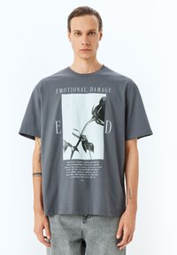 Camiseta de algodón gris con un gran gráfico de una rosa negra y el texto que dice "DAÑO EMOCIONAL", con mangas cortas y un corte relajado.