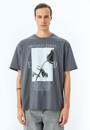 Camiseta de algodón gris con un gran gráfico de una rosa negra y el texto que dice "DAÑO EMOCIONAL", con mangas cortas y un corte relajado.