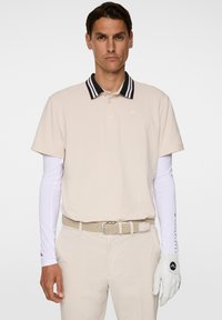 Beige polo skjorta med svart och vit randig krage, matchad med vita långärmade skjortor, beige byxor, flätad bälte och vit golfhandske.