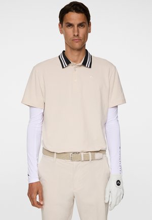 Beige polo-shirt med sort og hvidt stribet krave, parret med hvide langærmede skjorter, beige bukser, flettet bælte og hvid golfhandske.