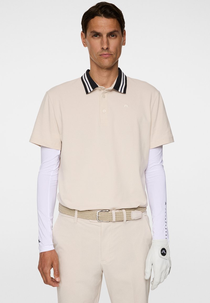 Beige polo skjorta med svart och vit randig krage, matchad med vita långärmade skjortor, beige byxor, flätad bälte och vit golfhandske.