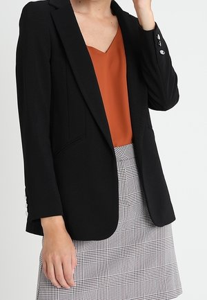 Femme portant un blazer noir sur un haut orange rouille à col en V et une jupe grise à carreaux, debout avec une main levée près du visage.