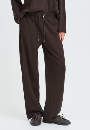 Personne portant un pantalon ample marron foncé avec une taille à cordon et des chaussures marron foncé à lacets, debout devant un fond clair uni.