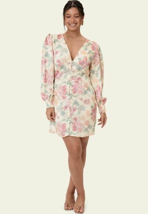 Femme souriante portant une robe fleurie à col en V avec des manches longues bouffantes et des boutons sur le devant, debout, jambes croisées, mains détendues.