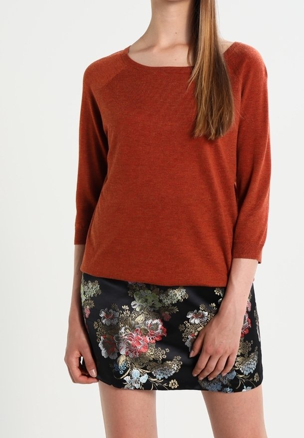 Femme portant un haut à manches longues couleur rouille et une mini-jupe noire à motif floral multicolore, debout devant un fond uni.