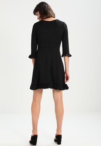 Robe noire avec un corsage ajusté et une jupe évasée. Manches trois-quarts avec des poignets à volants et un ourlet volanté. Texture lisse.