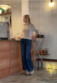 Maglione beige chiaro a maglia, jeans blu a gamba larga e calzature grigie. Bancone in legno con barattoli di vetro in un ambiente caffetteria minimalista.