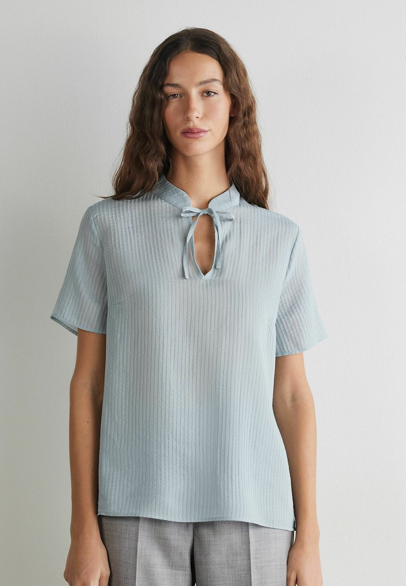 Woman with long wavy brown hair wears light blue ribbed short-sleeve top with keyhole neckline and tie, standing against plain wall.