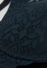 Tmavě modrá krajková podprsenka s květinovými vzory, texturovanou tkaninou a hladkým povrchem. Nastavitelné ramínka dotvářejí design.