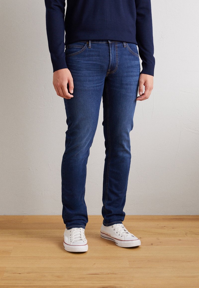 Jean slim bleu foncé en denim, doté d'un design à cinq poches et de coutures discrètes. Porté avec des baskets blanches sur un sol en bois.