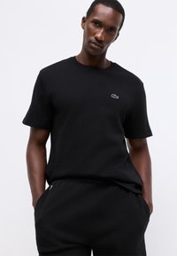 Homem vestido com uma t-shirt texturizada de manga curta preta e calças combinando, com as mãos nos bolsos contra um fundo simples.