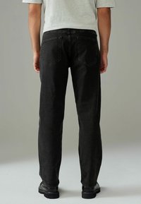 Zwarte denim broek met een rechte snit, voorzien van achterzakken en een gladde textuur, gedragen met een lichtgrijze shirt en zwarte schoenen.