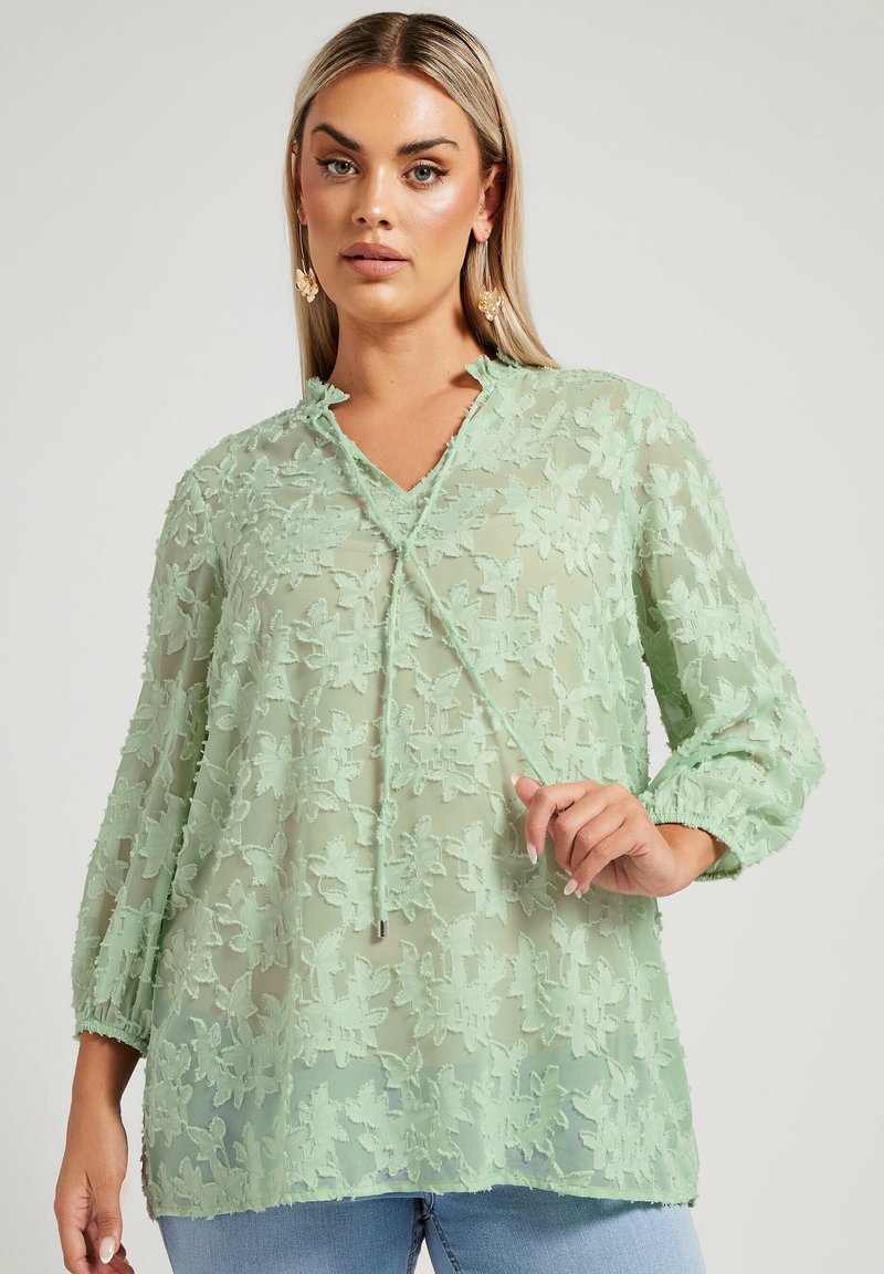Blusa verde menta realizzata in tessuto trasparente con ricami floreali, scollatura a V con lacci e maniche a tre quarti. Finitura testurizzata.