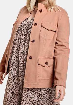 Kobieta ubrana w łososiową kurtkę na guziki z kieszeniami na klapę, na luźnej sukience w panterkę, stoi na tle jednolitego jasnego tła.