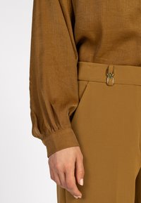 Close-up van een persoon in een bruine getextureerde blouse met lange mouwen en manchet, gecombineerd met bijpassende bruine high-waisted broek met gouden sluiting.