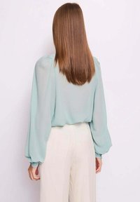 Blusa verde menta chiaro con maniche lunghe a sbuffo, tessuto trasparente e vestibilità morbida, abbinata a pantaloni crema a gamba larga.