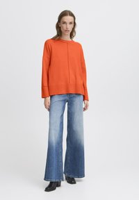 Nő áll egy élénk narancssárga, oversized pulóverben, világos-kék és sötétkék széles lábú nadrágban, fekete magassarkú bokacsizmában, sima háttér előtt.