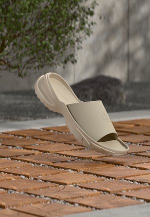 Μπεζ σανδάλι με χοντρή σόλα και ανοιχτή μύτη ξεκουράζεται πάνω σε ξύλινα πλακάκια με μικρές πέτρες ανάμεσα στα κενά, με θολό φόντο από πράσινο και τοίχο.