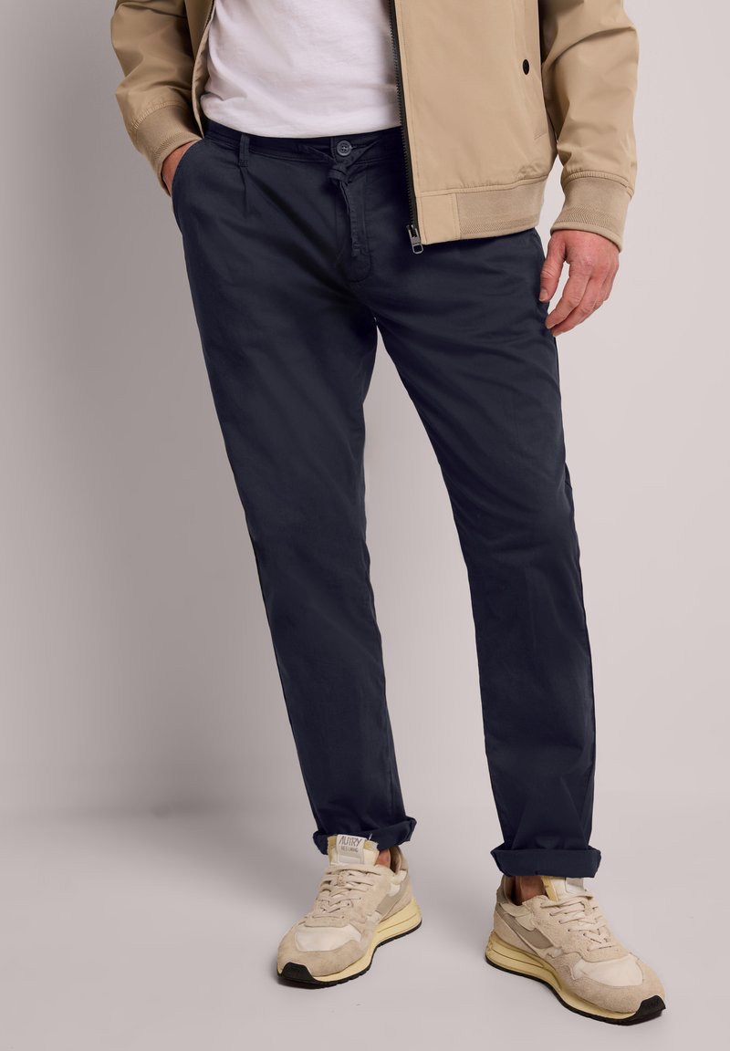 Homme portant une veste beige, un t-shirt blanc, un pantalon bleu marine et des baskets couleur crème, debout avec une main dans la poche.