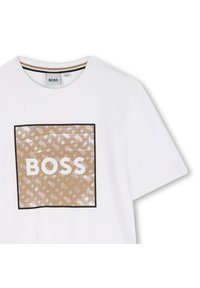 Bela bomba iz bombaža z velikim, vzorčastim kvadratom v bež barvi, na katerem je v debelih belih črkah v črnem okviru napisana "BOSS".