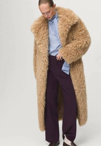 Femme portant un long manteau en fausse fourrure beige sur une blouse bleue avec un nœud et un pantalon large violet foncé, regardant vers le bas avec une main près de la taille.