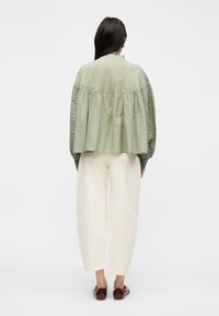 Γυναίκα με μακριά σκουρόχρωμα μαλλιά, φορώντας φαρδιά πουκάμισα πράσινου φασκόμηλου με υφασμάτινα μανίκια και κρεμ παντελόνια cropped, στέκεται γυρισμένη πλάτη πάνω σε λευκό φόντο.