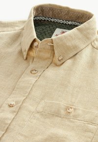 Camisa de botones beige claro con un tejido texturizado, que presenta un bolsillo, botones de aspecto perlado y un forro de cuello interior con un patrón.