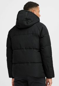 Sort pufferjakke med hætte, quiltet design og mat overflade. Funktioner inkluderer en løs pasform og fremtrædende sømme på skuldrene og ryggen.