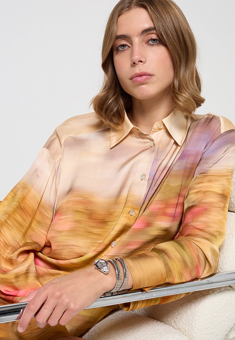 Seidenbluse mit buntem, abstraktem Farbverlauf in Orange-, Pink- und Violetttönen. Verfügt über einen Knopfverschluss und weite Ärmel. Silberne Uhr am Handgelenk.