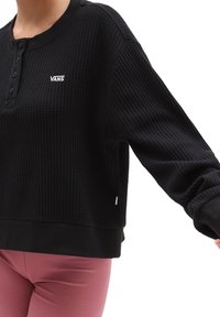 Zwarte wafelstructuur lange mouwen top met henley-halslijn en logo-detail. Heeft een cropped ontwerp en ribgebreide manchetten bij de polsen.