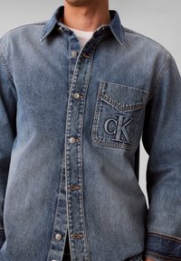 Homme portant une chemise en denim bleu à boutons avec une poche poitrine arborant le logo brodé "cK" et un t-shirt blanc visible en dessous.