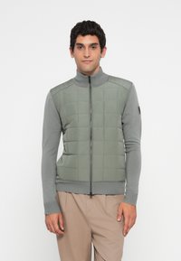 Man stands facing forward wearing a gray quilted zip-up jacket with knitted sleeves and beige pants against a plain white background.
