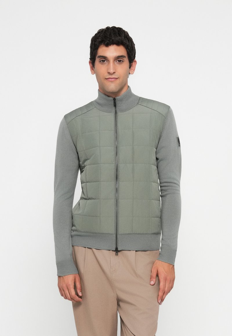 Man stands facing forward wearing a gray quilted zip-up jacket with knitted sleeves and beige pants against a plain white background.