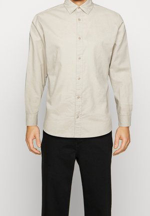 Mand iført en beige skjorte med knapper og lange ærmer samt sorte bukser, står afslappet med armene ned langs siden mod en ensfarvet baggrund.