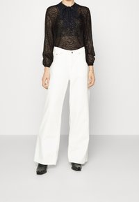 Blusa in pizzo nero con colletto a fiocco, abbinata a pantaloni bianchi a gamba larga e stivaletti neri alla caviglia. Texture trasparente sulla blusa.