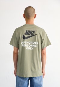 Olivinės spalvos medvilninis marškinėliai su juodu Nike logotipu ir tekstu "TIK ĮGALIOTI ASMENYS" ryškiomis baltomis raidėmis ant nugaros.