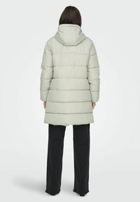 Manteau d'hiver léger gris clair avec une capuche, présentant un design matelassé et une coupe longueur genou. Porté avec un pantalon noir, vu de dos.