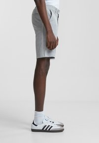 Shorts a cuadros grises con pliegues frontales y detalles de ribete blanco, combinados con zapatillas Adidas blancas, mostrados de perfil sobre un fondo neutro.