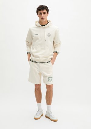 Νέος άνδρας με κρεμ φούτερ με κουκούλα με λογότυπο και ταιριαστό σορτς με αριθμό 67, στέκεται με τα χέρια στις τσέπες προς λευκό φόντο.