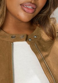 Brun mockajacka med tryckknappar och en central dragkedjedetalj, i kombination med en vit topp. Slät textur med en närbild av halsringningen.