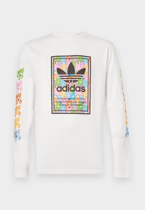 Νέος άνδρας που φοράει λευκό μακρυμάνικο Adidas μπλουζάκι με πολύχρωμο λουλουδένιο λογότυπο, μαύρη φαρδιά Adidas φόρμα με λευκές ρίγες, όρθιος.
