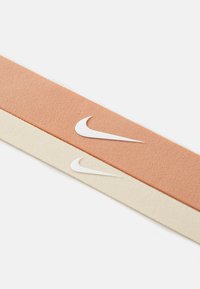Dva tekstilna remena, jedan breskvast i jedan kremasti, s bijelim Nike swoosh logotipima. Tekstura izgleda mekano s blagim rebrastim uzorkom.
