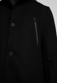 Schwarzer Wollmischungs-Mantel mit strukturiertem Finish, Knopfverschlüssen und einer Reißverschlusstasche auf der Brust in einem eleganten, minimalistischen Design.
