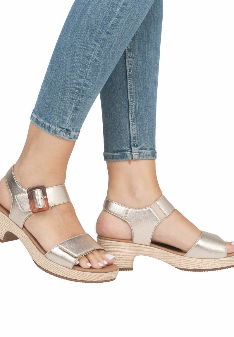 Sandales métalliques dorées avec un design à large bride, dotées d'une boucle sur le côté et d'un talon compensé texturé de couleur claire. Semelle extérieure en caoutchouc visible.