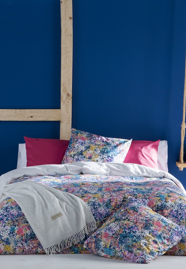 Cama con ropa de cama floral multicolor, dos almohadas rosas, una manta gris claro y un marco de madera rústico contra una pared azul oscuro.