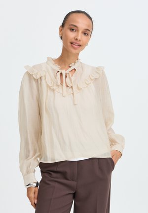 Vrouw draagt een licht beige blouse met ruches en lange mouwen en donkere bruine broek, glimlacht met één hand in haar zak, tegen een witte achtergrond.