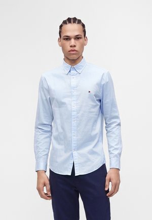 Jeune homme aux cheveux tressés portant une chemise bleu clair à boutons et un pantalon foncé, debout sur un fond blanc uni.