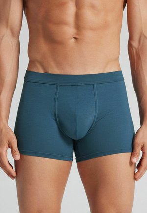 Männlicher Oberkörper mit türkisfarbenen Boxershorts, sichtbarem muskulösem Bauch und Armen vor einem einfarbigen hellen Hintergrund.